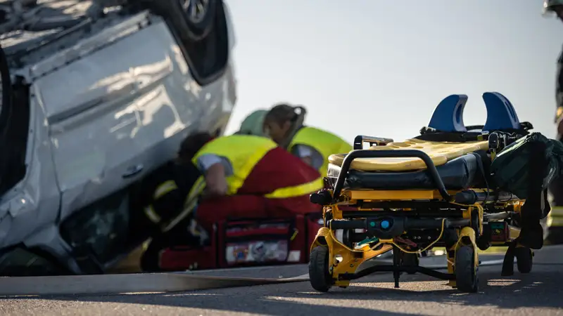 A gurney with first responders rescuing someone from an overturned car in the background